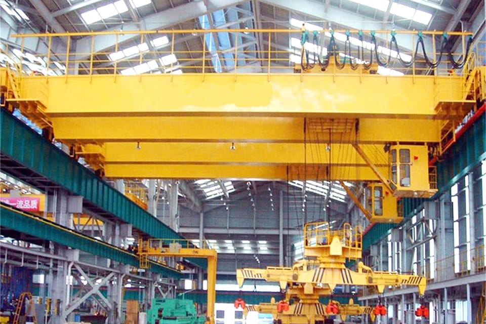 En traverskran med en betydelig gul drager udstyret med en kontrolkabine, der opererer inde i et industrilager med højt til loftet og flere arbejdsstationer.
