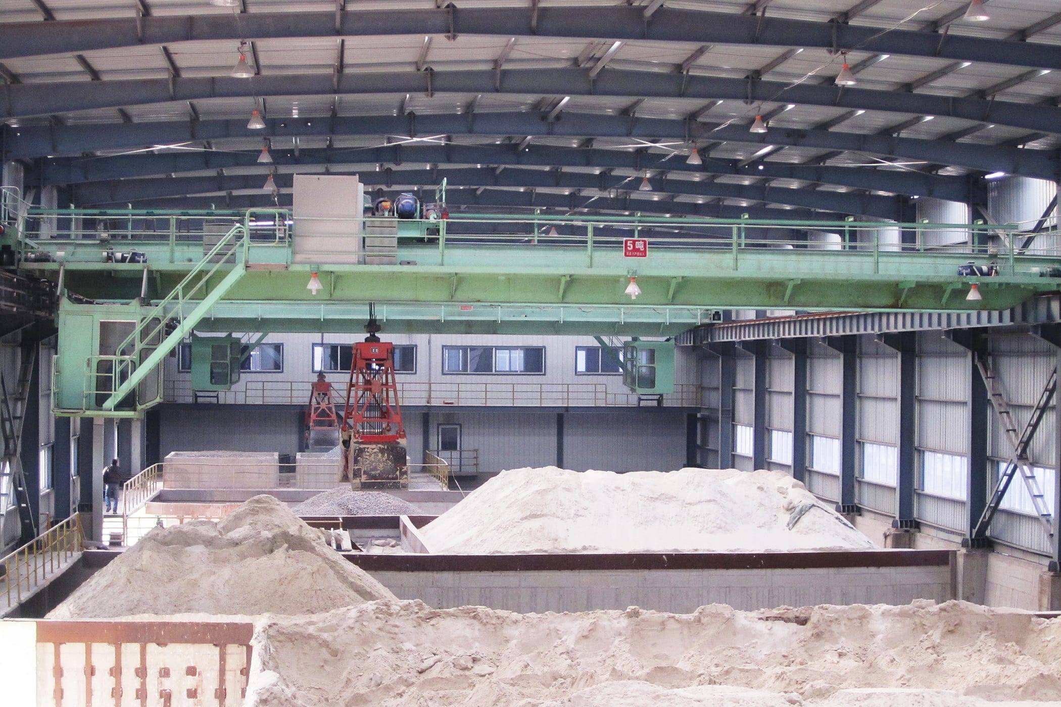 Een groene dubbelligger-grijper-bovenloopkraan in een industriële omgeving, gepositioneerd boven hopen grondstoffen, klaar voor de behandeling van bulkmateriaal