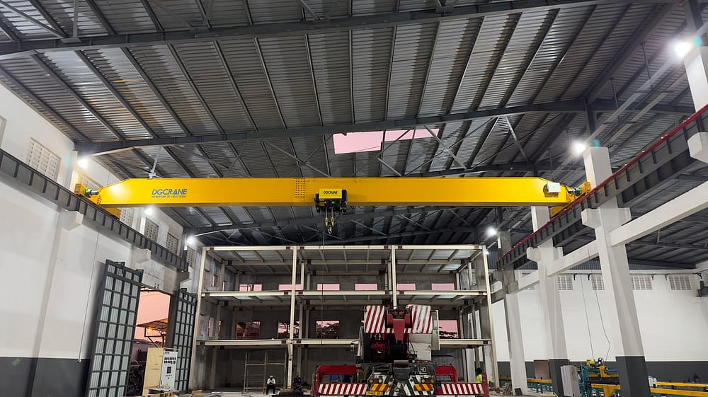 Żółta suwnica pomostowa FEM w standardzie FEM z brandingiem DGCRANE, zlokalizowana na dużym obszarze budownictwa przemysłowego z konstrukcjami metalowymi i ciężkimi maszynami poniżej
