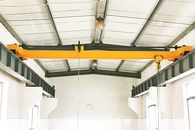  Een handmatige enkelligger bovenloopkraan met een gele balk en een hangende kettingtakel, geïnstalleerd in een wit industrieel gebouw.
