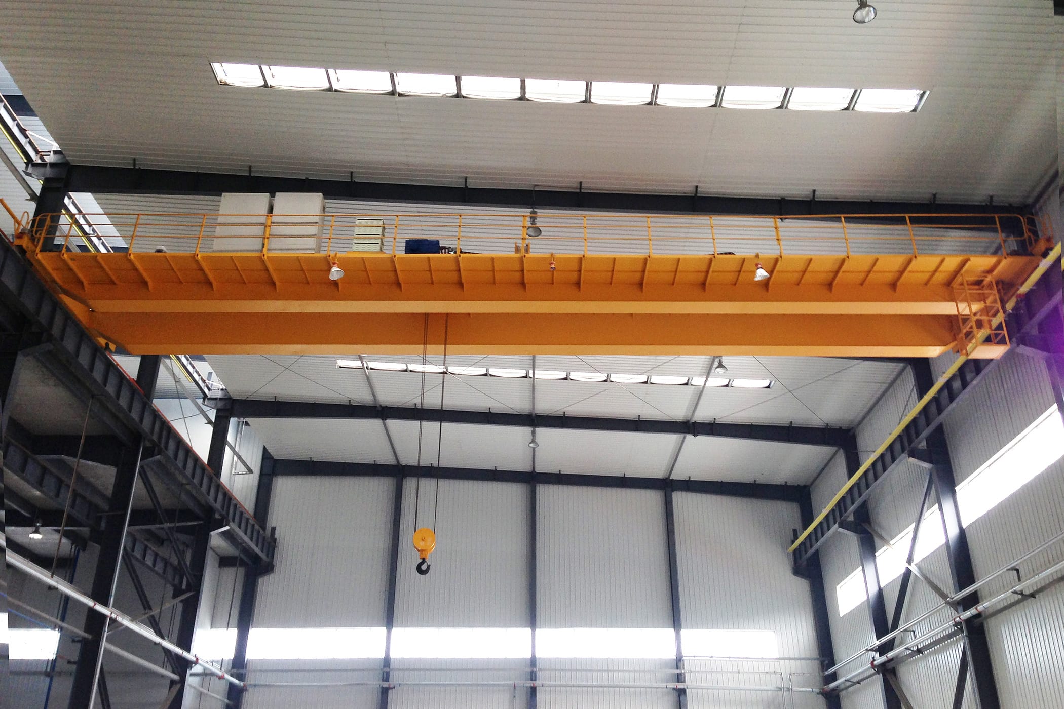 Een grote gele dubbelligger-bovenloopkraan van het type QD met een hangende oranje haak, die de breedte van een industrieel magazijn overspant met natuurlijk licht dat door dakramen in het dak komt