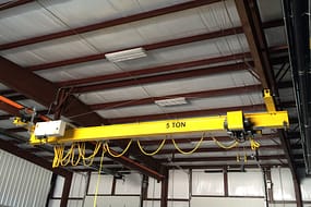  A bright yellow low headroom single girder overhead crane, with 5 TON prominently displayed, hanging from a red steel structure in a warehouse with white insulated paneling