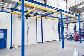  Een bovenloopkraansysteem voor werkstations met blauwstalen steunkolommen en een gele balk, voorzien van een takel, in een schone en georganiseerde industriële omgeving